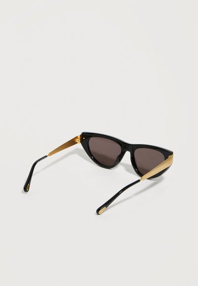 Μαύρα γυαλιά ηλίου σε σχήμα γατίσιου ματιού με σκούρους φακούς και χρυσά μεταλλικά μπράτσα που ακουμπούν σε φωτεινή επιφάνεια.