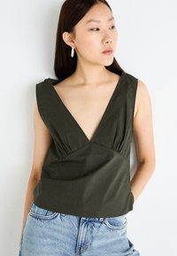 Dark green sleeveless top with a deep V-neckline, gathered detailing at the waist, and a smooth texture, paired with light blue jeans.