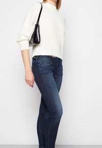 Žena v bílém pleteném svetru a tmavě modrých skinny džínách, nesoucí malou černou kabelku přes rameno na jednolitém světlém pozadí.