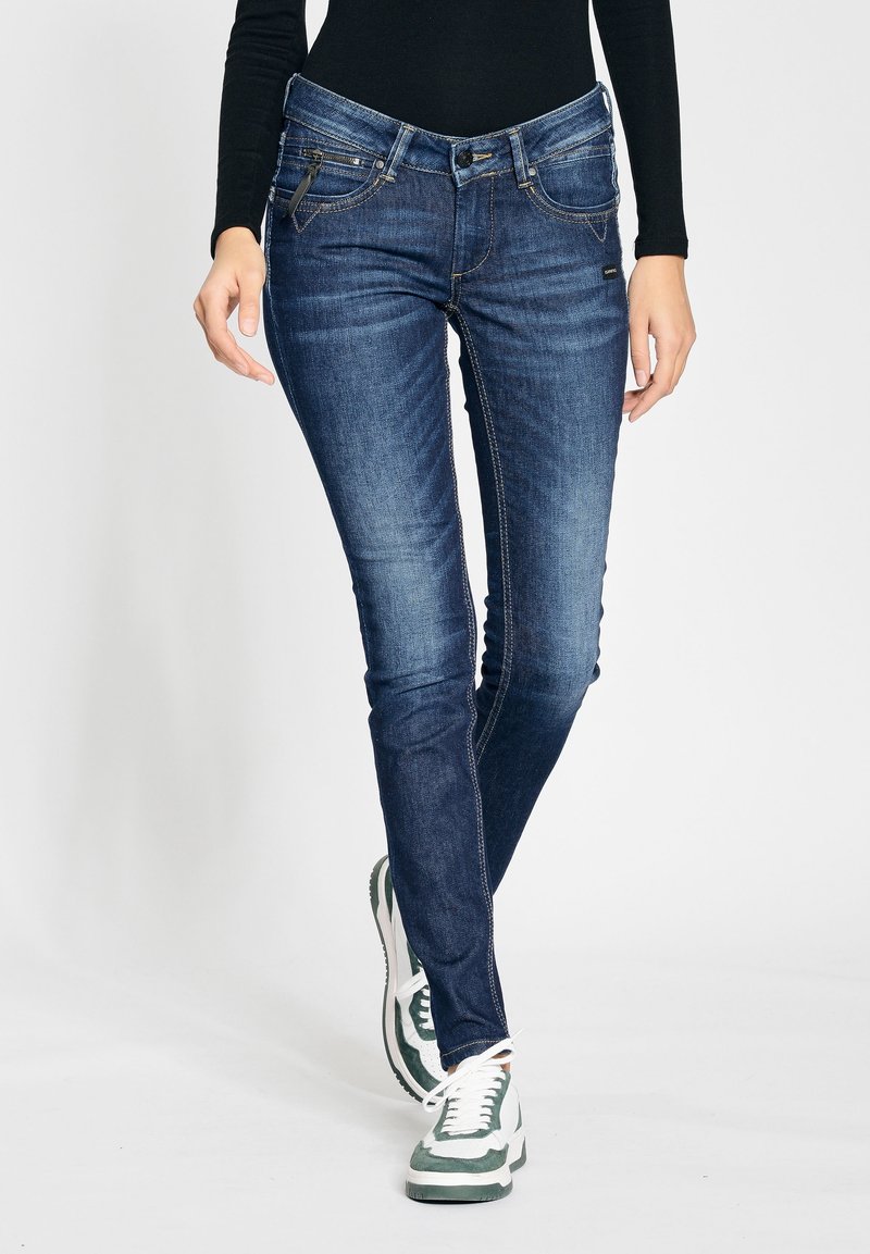Skinny-fit denim jeans in dark blue with subtle fading, five pockets, and contrasting stitching. Worn with green and white sneakers.