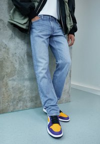 Person som bär ljusblå jeans, svart och vit jacka, samt skor i stark orange, lila och vitt, står mot en betongvägg.