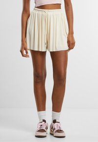 Ljusgräddefärgade shorts med elastisk midja, plisserad design och dragsko. Matchas med rosa och bruna sneakers och vita ankelsockor.