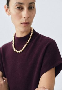 Bordeaux wollen top met een hoge halslijn en korte mouwen, gecombineerd met een gouden kralenketting, benadrukt de gladde texturen en het klassieke ontwerp.