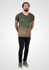 Kortærmet olivengrøn og brun farveblokeret t-shirt med en lomme foran, parret med sorte jeans og olivengrønne sneakers.