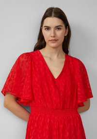 Jonge vrouw met lang bruin haar, gekleed in een rode jurk met doorzichtige, vloeiende mouwen en een V-halslijn, tegen een effen lichtgrijze achtergrond.