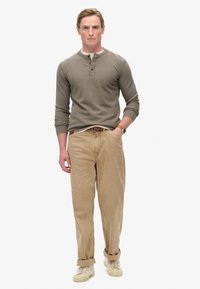 Olijfgroene henley-shirt met lange mouwen en knoopsluiting, gecombineerd met beige broek en crèmekleurige sneakers. Eenvoudig ontwerp en relaxte pasvorm.