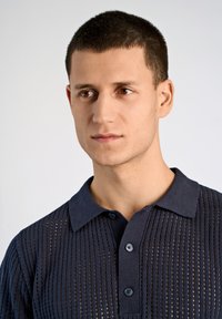 Marinblå stickad polotröja med en strukturerad öppen väv, utrustad med flat krage och treknappsslå, accentuerad av mörka knappar.