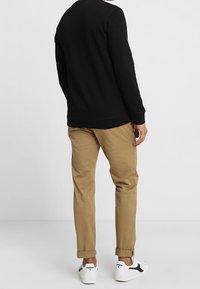 Mężczyzna w czarnym swetrze, khaki spodniach i białych trampkach stoi tyłem do kamery na jednolitym białym tle.