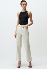 Zwarte geribbelde tanktop gecombineerd met hooggepareerde, wijde lichtbeige broek. De outfit heeft twee voorzakken en een knoopsluiting.