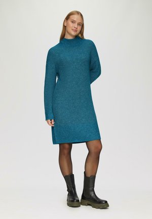 Robe pull en tricot teal avec un col montant et un ourlet côtelé. Dotée de manches longues et d'un tissu texturé. Associée à des bottines noires à semelle épaisse.