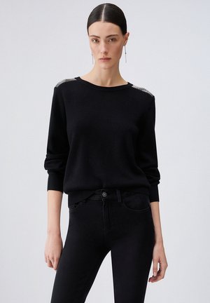Donna con capelli tirati indietro che indossa un maglione nero con dettagli a quadri sulle spalle e pantaloni neri, in piedi davanti a uno sfondo semplice.