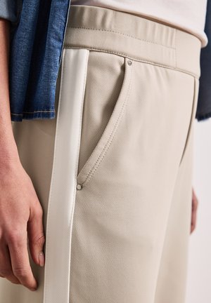 Beige broek met witte zijstreep en detail op de voorzak, gedragen met een licht topje ingestopt en een blauwe spijkerjas over de arm.