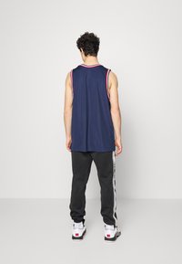 Laivastonsininen hihaton urheilutoppi, jossa on punaiset ja valkoiset reunukset, yhdistetty harmaisiin vetoketjullisiin joggereihin, joissa on sivuraidat, ja valkoiset tennarit jalassa.