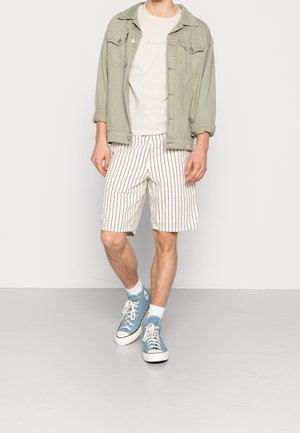 Lichtgroene denimjas over een beige T-shirt, gecombineerd met beige gestreepte shorts en blauwe hoge sneakers met witte zolen en sokken.