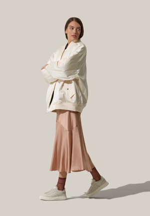 Cremfarbener Bomberjacke über einem dunklen Oberteil, kombiniert mit einem fließenden rosa Midirock und beigen Sneakers; bietet glatte Texturen und geschichtete Designs.
