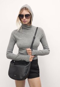 Maglione a collo alto grigio a costine con maniche lunghe, abbinato a una borsa a tracolla in pelle nera. Il modello indossa occhiali da sole oversize neri.