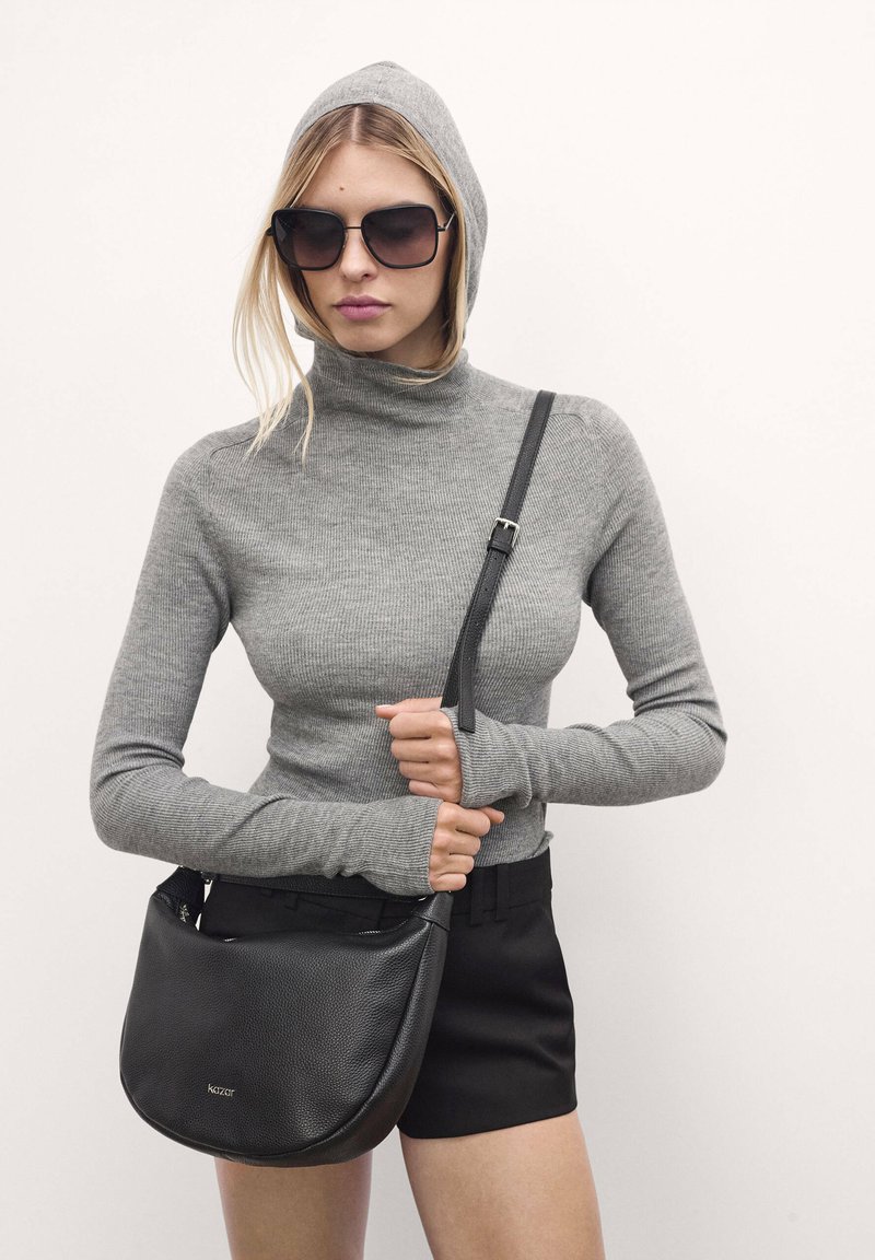 Maglione a collo alto grigio a costine con maniche lunghe, abbinato a una borsa a tracolla in pelle nera. Il modello indossa occhiali da sole oversize neri.