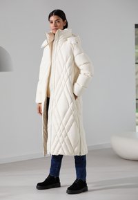 Long, quilted coat in cream with a high collar, front zipper closure, and drop shoulders. Worn over dark jeans and black ankle boots.