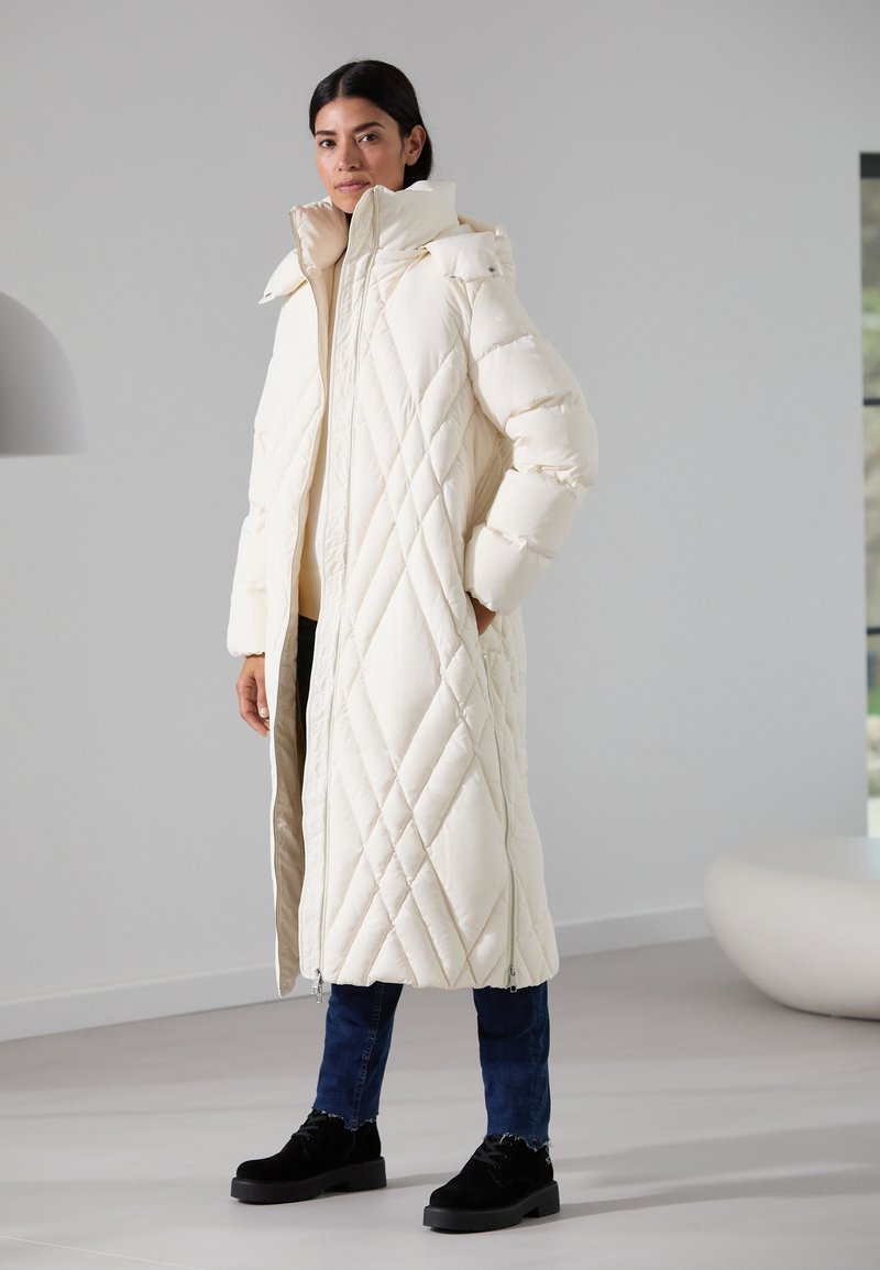 Long, quilted coat in cream with a high collar, front zipper closure, and drop shoulders. Worn over dark jeans and black ankle boots.