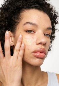 Modèle appliquant une crème beige clair sur sa joue, avec des cheveux noirs bouclés et une peau lisse avec des imperfections visibles. Elle porte des boucles d'oreilles en argent.