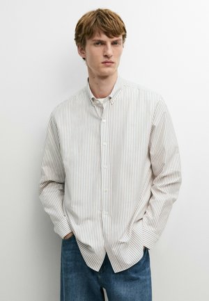 Jeune homme portant une chemise blanche à fines rayures verticales et un jean bleu, debout avec les mains dans les poches devant un fond blanc.