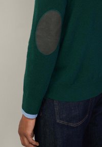 Vihreä neulepaita, jossa on tummanharmaa kyynärpäätuki, ja vaaleansininen hiha pilkistää alta, yhdistettynä tummiin farkkuihin.