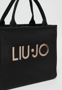 Borsa tote in nylon nera con doppi manici superiori e un logo metallico dorato "LIU·JO" ben visibile sul fronte. Texture liscia e forma strutturata.