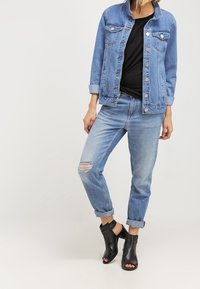 Žena v manžetových světle modrých roztrhaných džínách, riflové bundě přes černý top a černých sandálech na odkryté patě, stojící na jednoduchém pozadí.