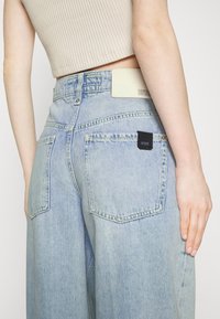 Vaaleansiniset farkut, joissa on korkeavyötärö ja leveät lahkeet, sekä kaksi takataskua. Materiaali on texturne, ja vyötärönauhassa on brändilogo.