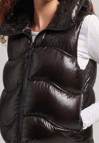 Czarna, pikowana kamizelka z błyszczącą teksturą, wysokim kołnierzem i pełnym zamkiem. Poziome szwy tworzą wzór przypominający pikowanie z wyraźnie zdefiniowanymi sekcjami.