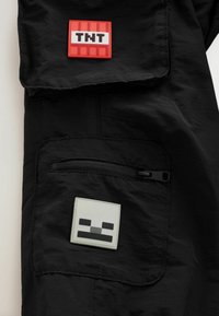 Fekete szövetkabát, két felvarróval: egy piros "TNT" felvarró és egy téglalap alakú szürke felvarró, pixelált arccal. Cipzáras zseb részletek.