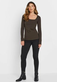 Langärmliges, dunkelbraunes, eng anliegendes Oberteil mit einem quadratischen Ausschnitt, kombiniert mit schwarzen engen Jeans und Stiefeletten. Glatte Textur und Silhouette.