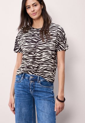 Frau trägt ein schwarz-weißes kurzärmeliges Shirt mit Zebramuster, blaue Jeans und ein dunkles Armband, steht vor einem einfarbigen hellen Hintergrund.