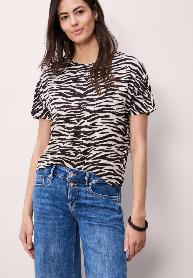 Frau trägt ein schwarz-weißes kurzärmeliges Shirt mit Zebramuster, blaue Jeans und ein dunkles Armband, steht vor einem einfarbigen hellen Hintergrund.