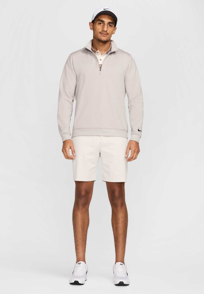 Helle beige Halbzip-Pullover mit einem kontrastierenden Kragen, kombiniert mit cremefarbenen Shorts. Die Accessoires bestehen aus einer weißen Nike-Cap und weißen Turnschuhen.