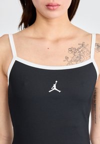 Zwarte geribbelde tanktop met witte schouders en een klein wit Jumpman-logo in het midden. Gladde textuur, atletische pasvorm.