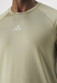 Camiseta atlética de manga corta en verde oliva, hecha de una tela suave y ligera. Presenta un sutil logo plateado de Adidas en el pecho.
