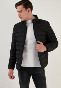Jachetă puf din neopren negru, cu linii orizontale matlasate, guler înalt și fermoar frontal. Purtată deasupra unei pulovere albe texturate și a unor blugi negri.