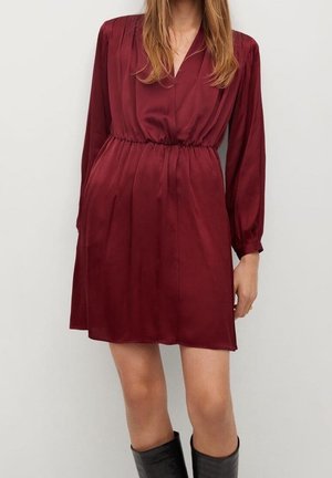 Kobieta w długiej, do kolan, głęboko czerwonej satynowej sukience z wcięciem w talii i czarnych, sięgających do kolan botkach, na tle jednolitego jasnego tła.