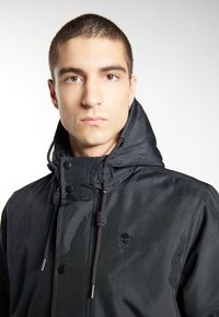 Schwarze Jacke mit Kapuze, die ein dezentes Totenkopf-Logo, eine Kordelzugverschluss und einen glatten, strukturierten Stoff aufweist.