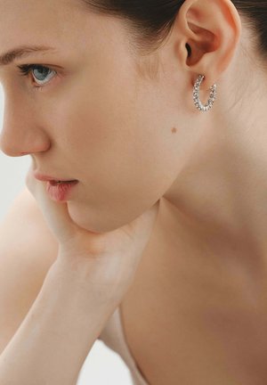 Joven mujer apoyando la barbilla en su mano, lleva un pendiente de aro plateado con pequeñas piedras brillantes.