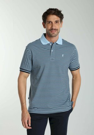 Homme aux cheveux courts et foncés portant un polo rayé bleu clair et noir, debout avec une main dans la poche sur fond blanc.