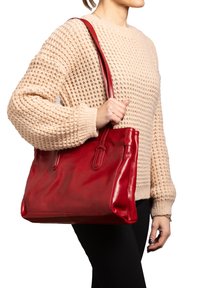 Rode leren tote bag met een gladde textuur, dubbele handvatten en subtiele details aan de zijkanten. Model draagt een beige gebreide trui en zwarte broek.