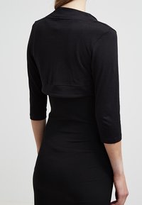 Avskuren svart jacka i mjukt tyg med trekvartslånga ärmar och rundad krage, matchad med en kroppsnära svart klänning.