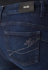 Džínové kalhoty v tmavě modré barvě s hladkou texturou, přední kapsa s designem ozdobeným malými kovovými nýty v květinovém vzoru.