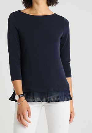 Femme portant un haut bleu marine à manches longues avec un détail d'ourlet plissé et un pantalon blanc, accessoirisée d'une montre et d'un bracelet.