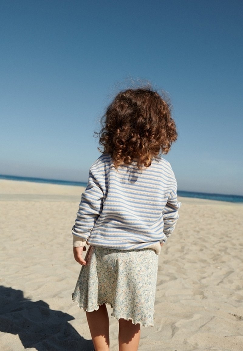 Enfant aux cheveux bouclés portant un pull rayé et une jupe fleurie, debout sur une plage de sable, face à une mer calme sous un ciel bleu clair.