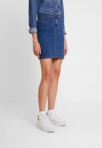 Denim Bleistiftrock in dunkelblau, mit schlankem Schnitt und vorderen Taschen. Getragen mit weißen Turnschuhen und weißen Knöchelsocken. Glatte Textur.