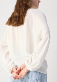 Witte, lichtgewicht blouse met uitlopende mouwen, zachte textuur en elastische manchetten. Gekleed met lichtblauwe denim broeken, handen samengevouwen achter de rug.