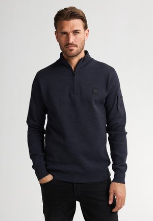 Man met lichtbruin haar, gekleed in een marineblauwe trui met een kwart rits, een borstembleem en een zwarte broek, staand tegen een lichte achtergrond.